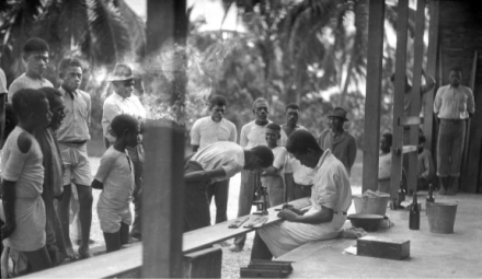 <p>Physician Malakai Veisamasama (seated) of Fiji invites Ni-Vanuatu to examine a hookworm sample, c. 1925<br />
Malakula, Vanuatu<br />
Photograph by Sylvester Maxwell Lambert</p>
<p>Public health physician Sylvester Maxwell Lambert, who worked for the International Health Division of the Rockefeller Foundation, traveled extensively between 1919 and 1939, visiting Kiribati, Papua New Guinea, the Solomon Islands, Tonga, Tuvalu, and Vanuatu. His focus was to study hookworm disease in hopes of eradicating it. Lambert also advocated for training local students to become doctors, such as Malakai Veisamasama, seen here.</p>
<p>University of California, San Diego Archives, bb1639202z_1</p>
